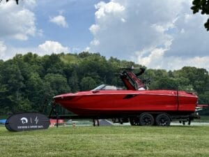 A red Centurion & Supreme boat is displayed on a trailer at a lakeside location, with trees and cloudy sky in the background. A black and white promotional sign stands nearby on the grass.