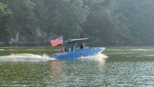 A blue motorboat with an American flag navigates on a calm green water lake; two people are visible on the boat, and the background consists of a forested shoreline.