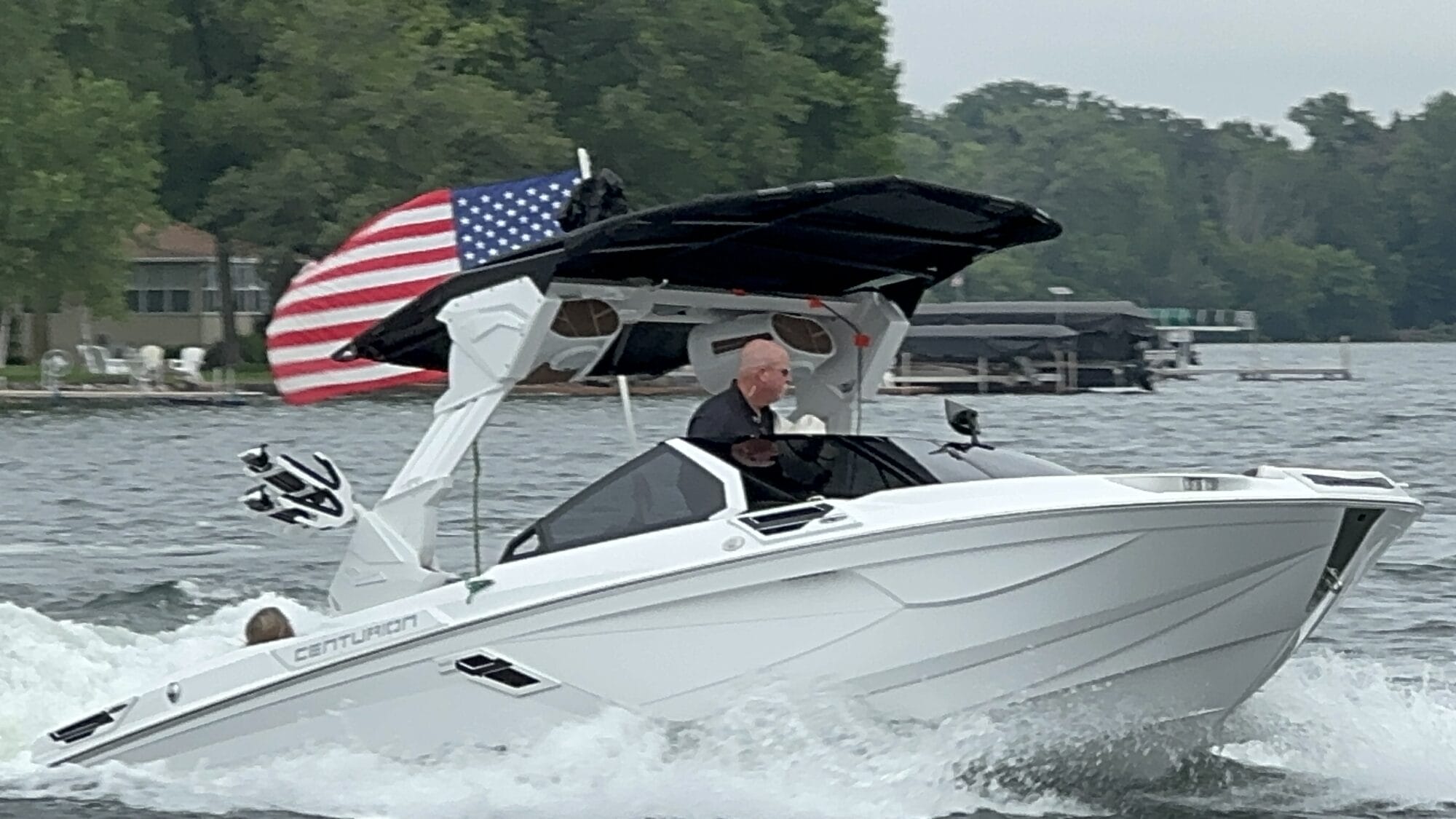 A person is driving a white Centurion boat on a lake. The boat has an American flag at the back, and there are trees and houses visible in the background.