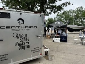 Centurion Boats promotional setup featuring branded trailer, merchandise stand, and tent with staff. A wake surfing boat is displayed in the background.