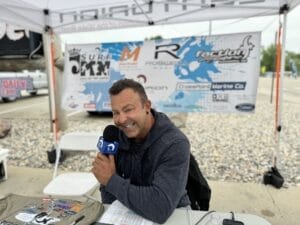 A man sitting at a booth, holding a microphone and smiling, with banners and a table in front of him. The table has papers and other items, and the background shows a vehicle and outdoor setting.