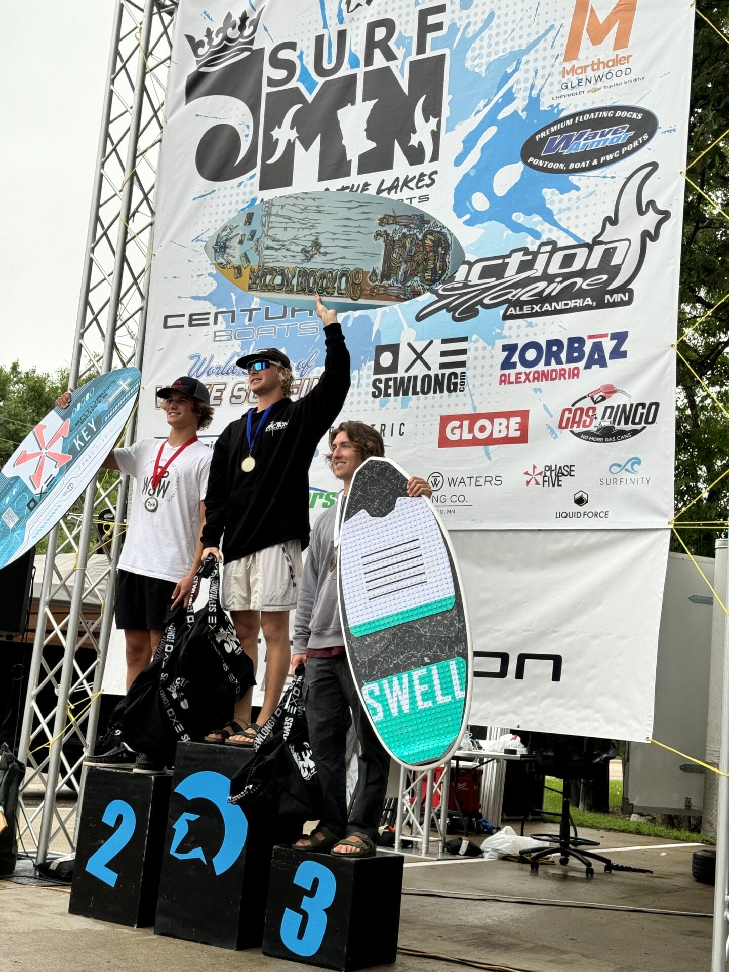 Three individuals stand on a podium holding awards at a surfing competition. The backdrop features various sponsor logos and a banner that reads "Surf MN." Two of them hold surfboards.