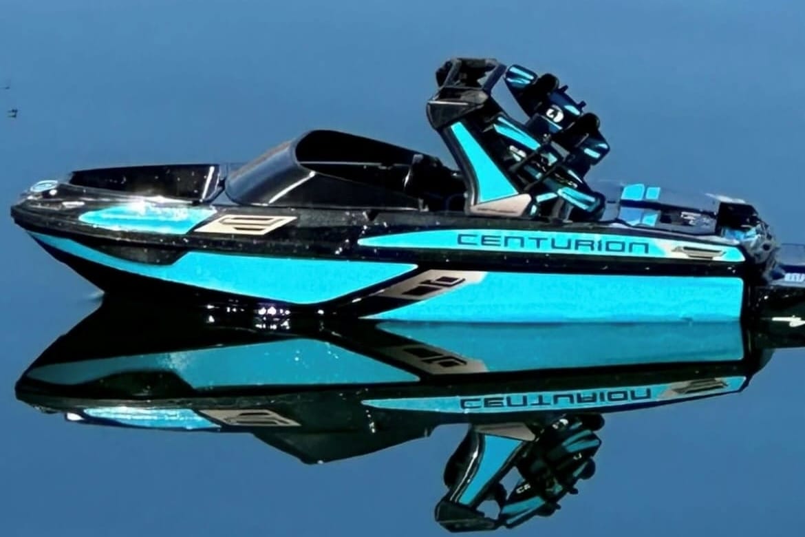 A blue Centurion wakeboard boat floats on calm water, perfectly reflecting its image.