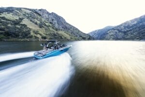 A blue motorboat speeds across a narrow river or lake, surrounded by hills and rocky terrain under a bright sky.