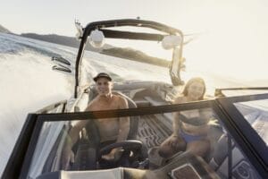 A man and a woman are enjoying a boat ride under the bright sun. The man is steering the boat, and both are smiling. Water is splashing in the background.