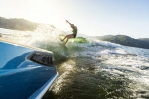 Person wakeboarding on a sunny day, performing a trick on the wake created by a boat in a lake surrounded by hills.