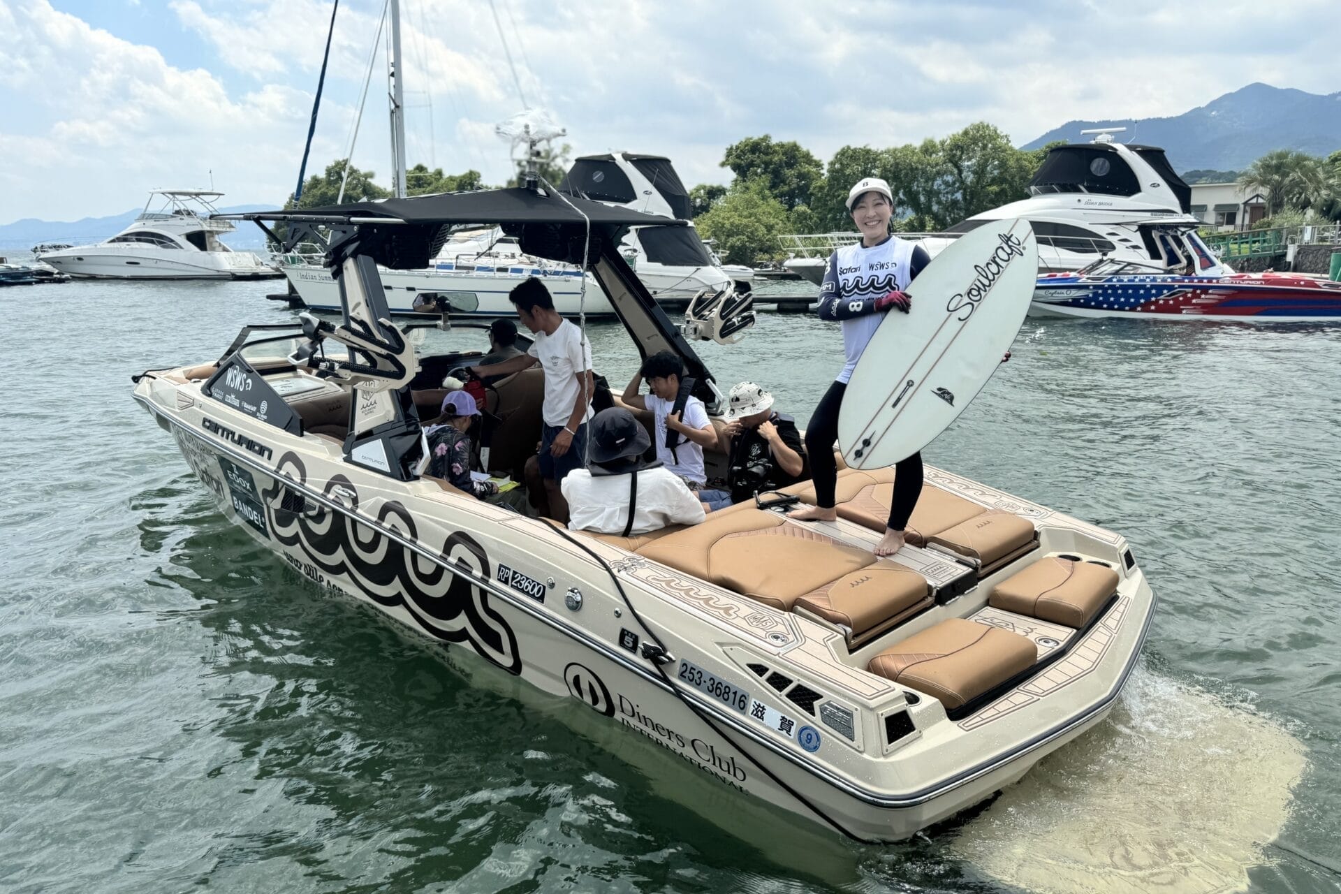 Group of people on a motorboat at a marina, one person standing with a surfboard. Other boats are visible in the background.