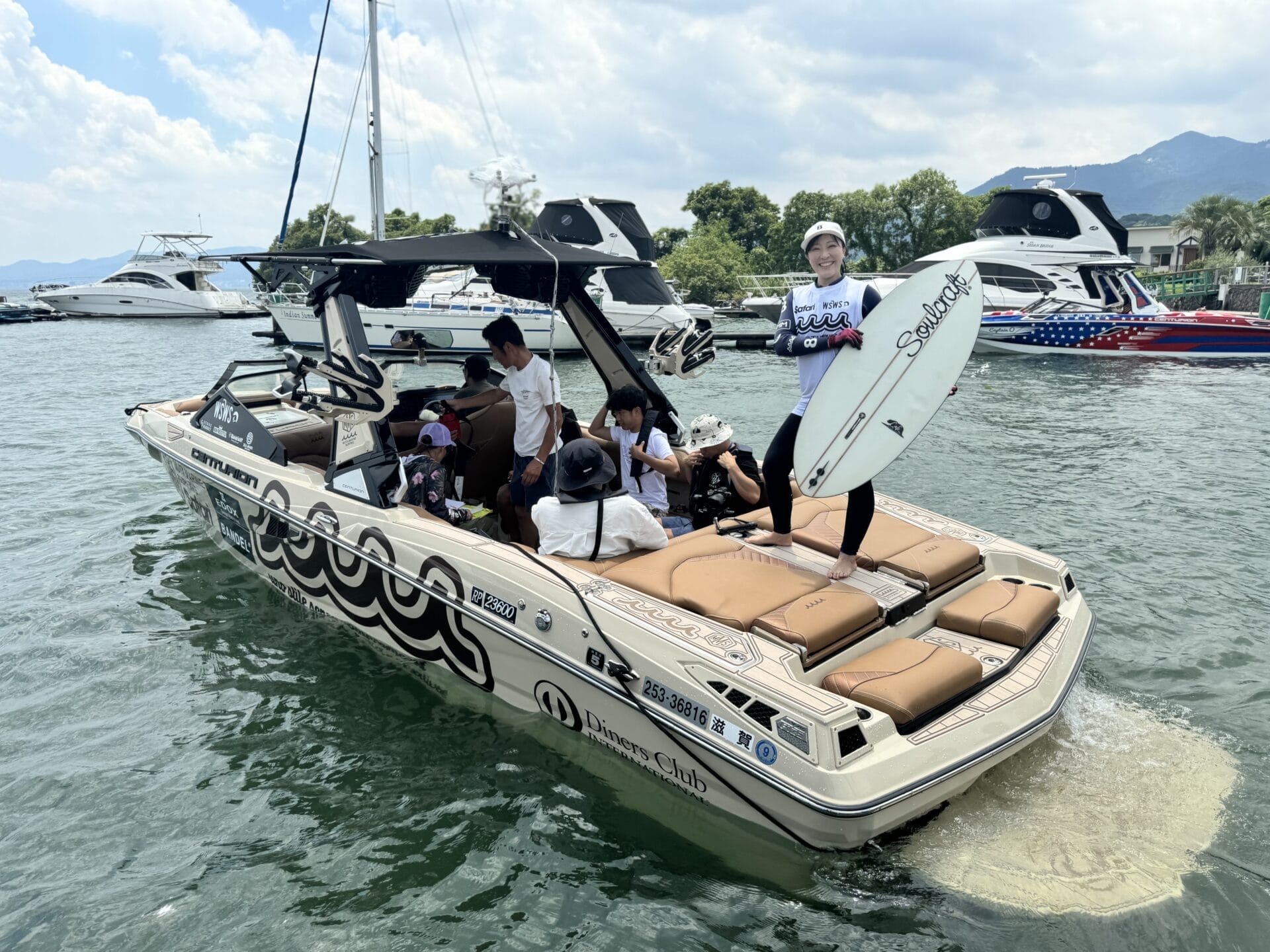 Group of people on a motorboat at a marina, one person standing with a surfboard. Other boats are visible in the background.