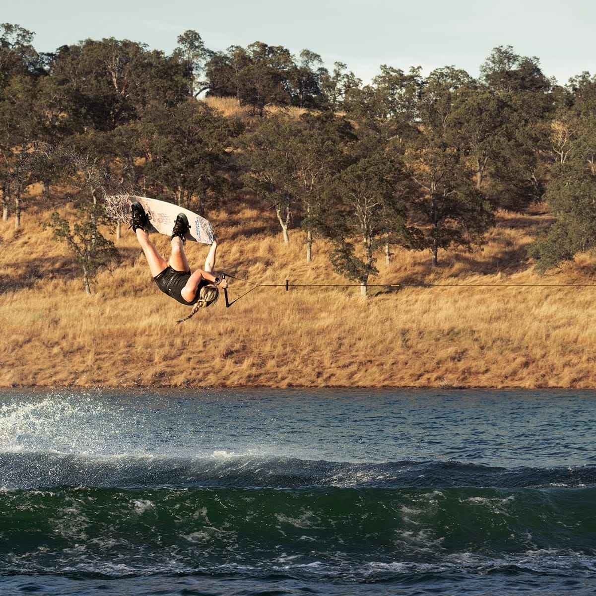 A person is wakeboarding in mid-air performing a flip above the water at a lake with trees and hills in the background.