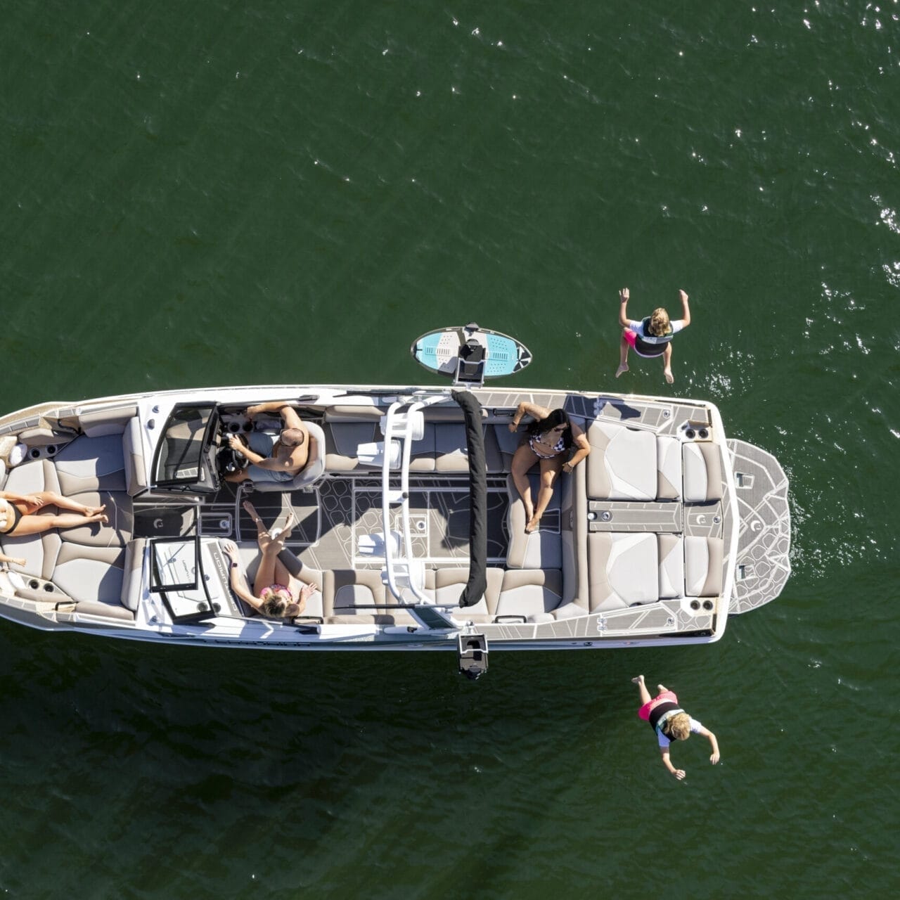 Aerial view of a boat on water with several people onboard. One person is lying down sunbathing, another is steering, and two individuals are in the water near the boat.