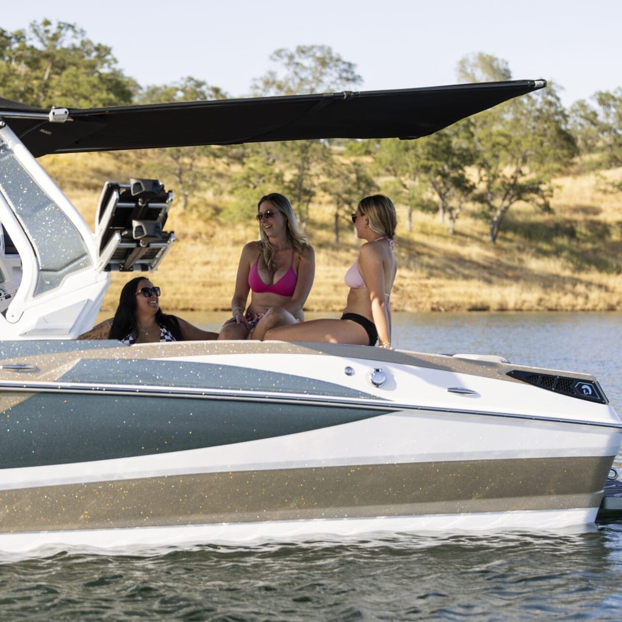 Three people are sitting and chatting on a boat in a body of water, surrounded by a natural landscape with trees and dry grass.