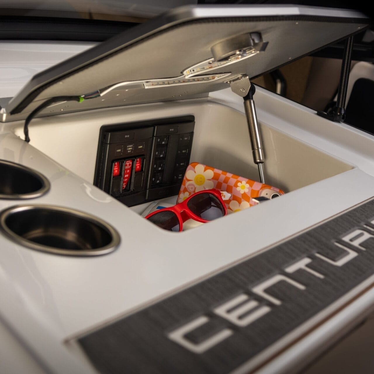 Open storage compartment on a Centurion boat, containing a pair of red sunglasses, a floral-patterned item, and a control panel with switches. Two cup holders are visible beside the compartment.