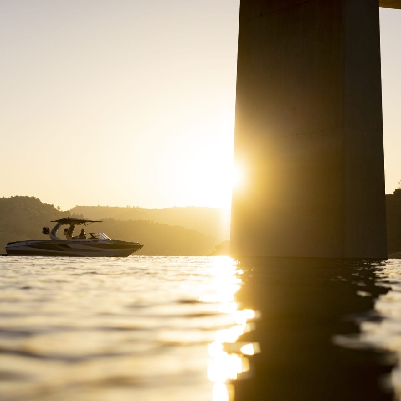 A boat is on the water near a bridge pillar during sunset. The sky is clear, and the sun is partially obscured by the pillar. The landscape in the background is hilly.