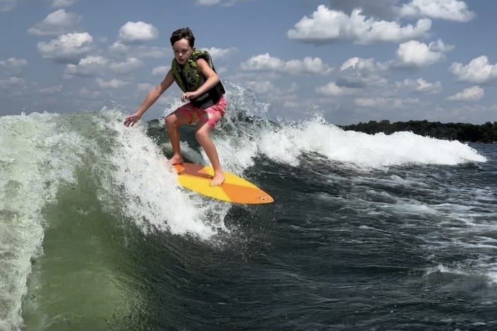 A person wearing a life vest rides a yellow surfboard on a wave under a blue sky with scattered clouds.