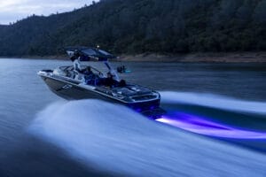 A speedboat with blue underglow lights moves swiftly across the water with a forested coastline in the background.