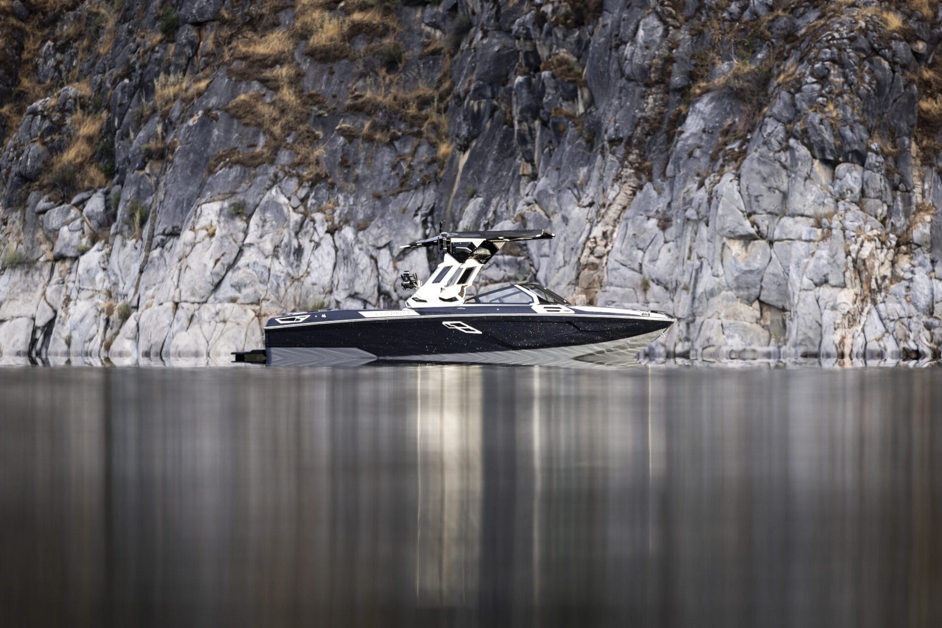 A black and white speedboat is anchored near a rocky cliff shoreline, partially reflected in the calm water.