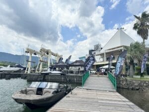 A docked boat next to a green-brightened wooden pier, with an event setup onshore featuring flags and tents from various brands and a building in the background under a cloudy sky.