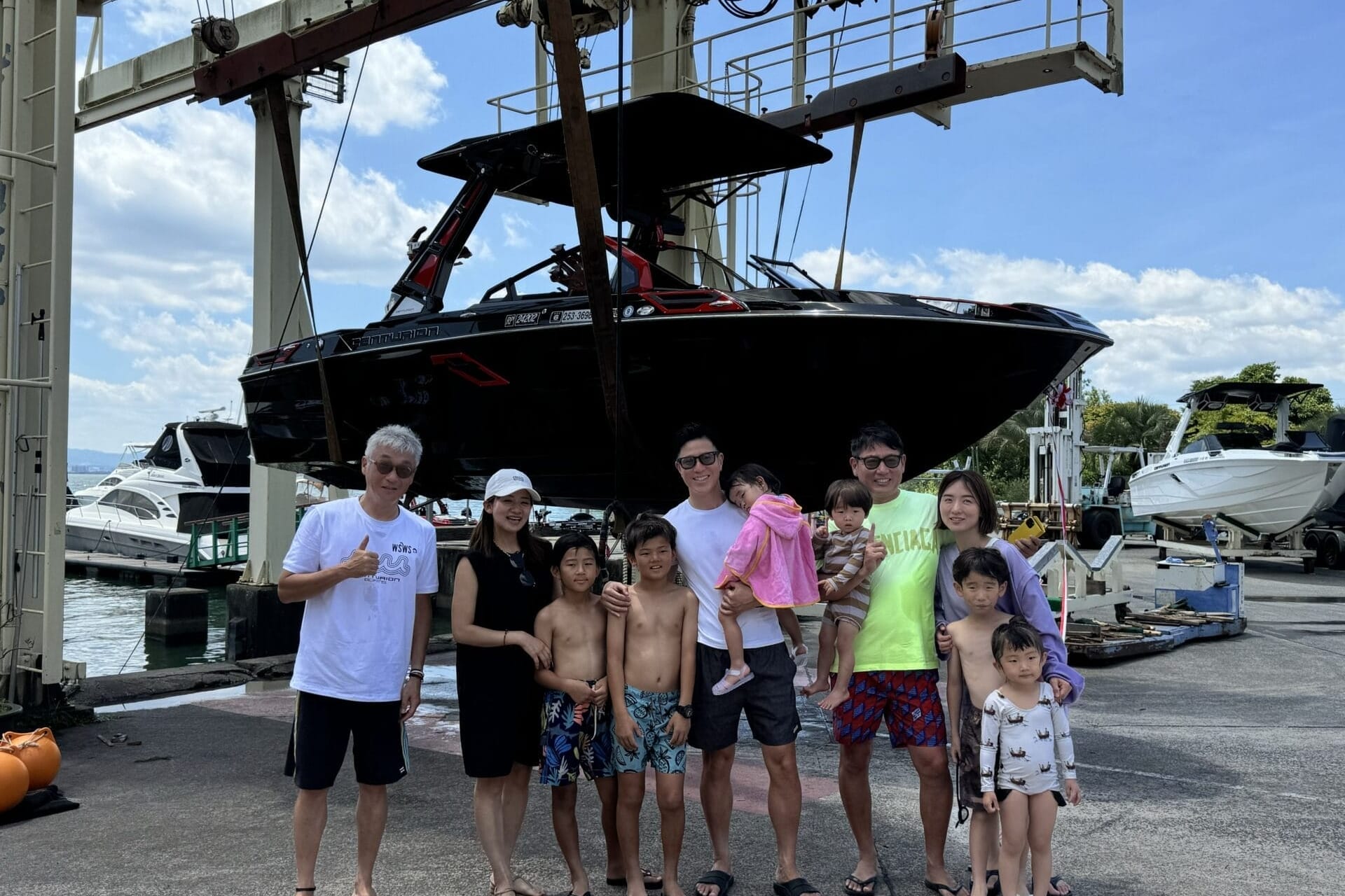 A group of people, including men, women, and children, stand together in front of a large boat suspended in the air by a mechanical lift at a marina.