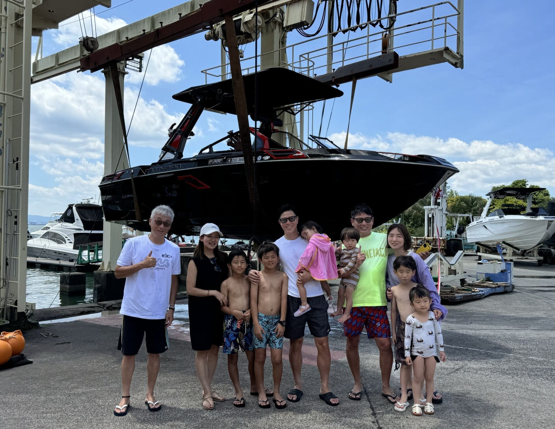 A group of people, including men, women, and children, stand together in front of a large boat suspended in the air by a mechanical lift at a marina.