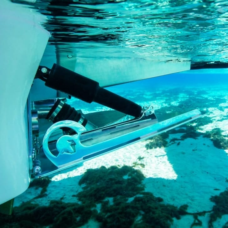 A metallic marine device is partially submerged in clear turquoise water, attached to a white surface. The device includes hydraulic components and is positioned above a sandy seabed with some seaweed.