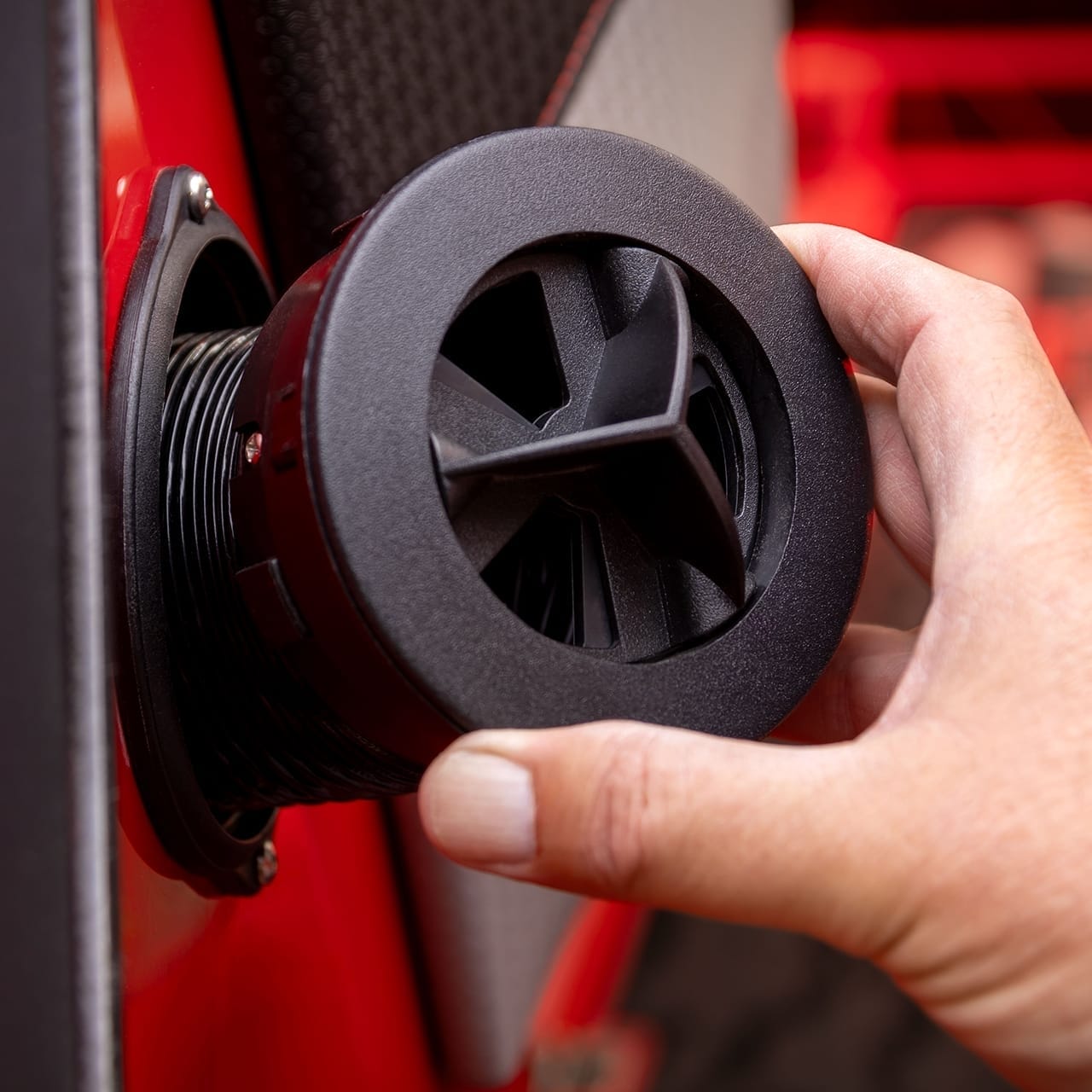 A hand is adjusting a black, circular car interior vent mounted on a red and gray dashboard.