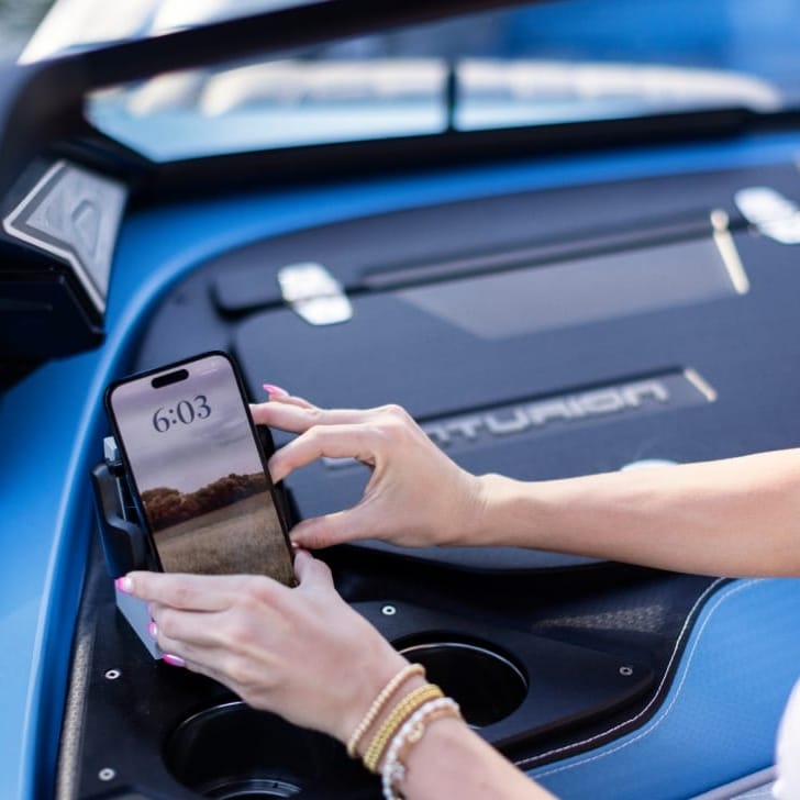 A person places a smartphone into a holder on a boat dashboard. The phone's screen shows the time as 6:03.