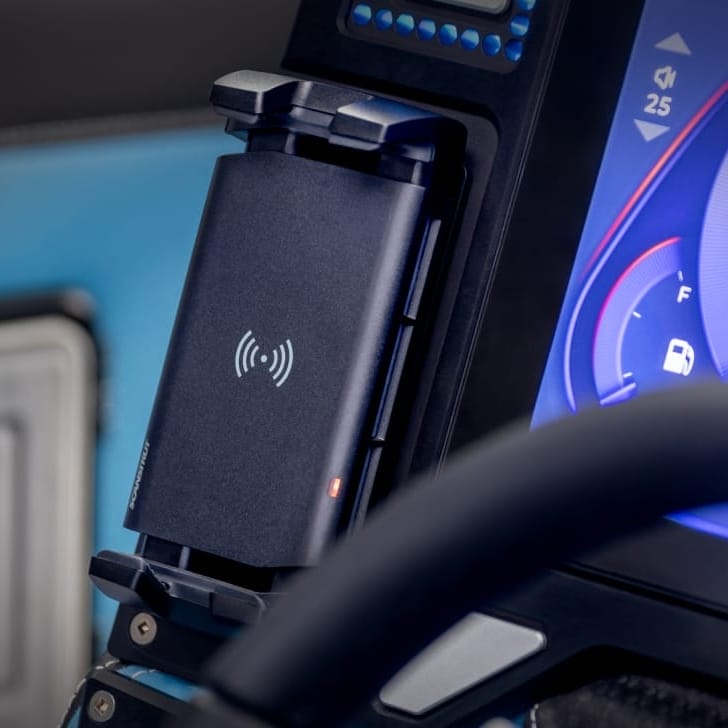 A black wireless charging phone mount is installed on a car dashboard next to the steering wheel, with a digital speedometer display in the background.