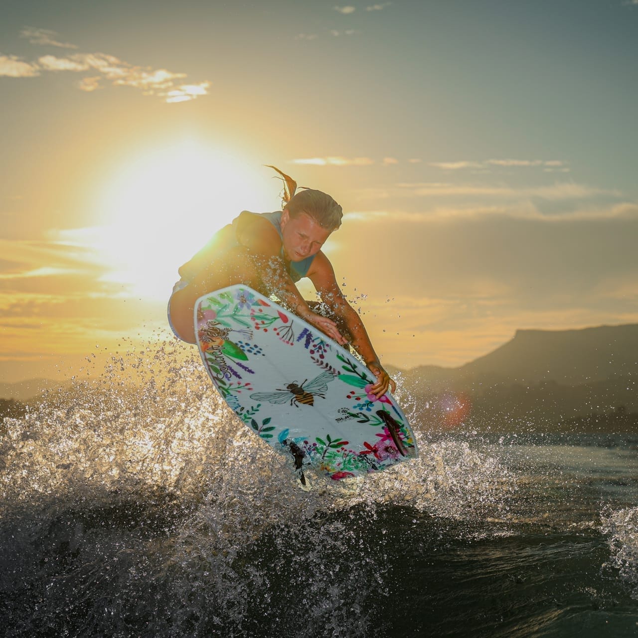 A person surfing on a wave during sunset, performing an aerial trick with a surfboard decorated with a colorful pattern.