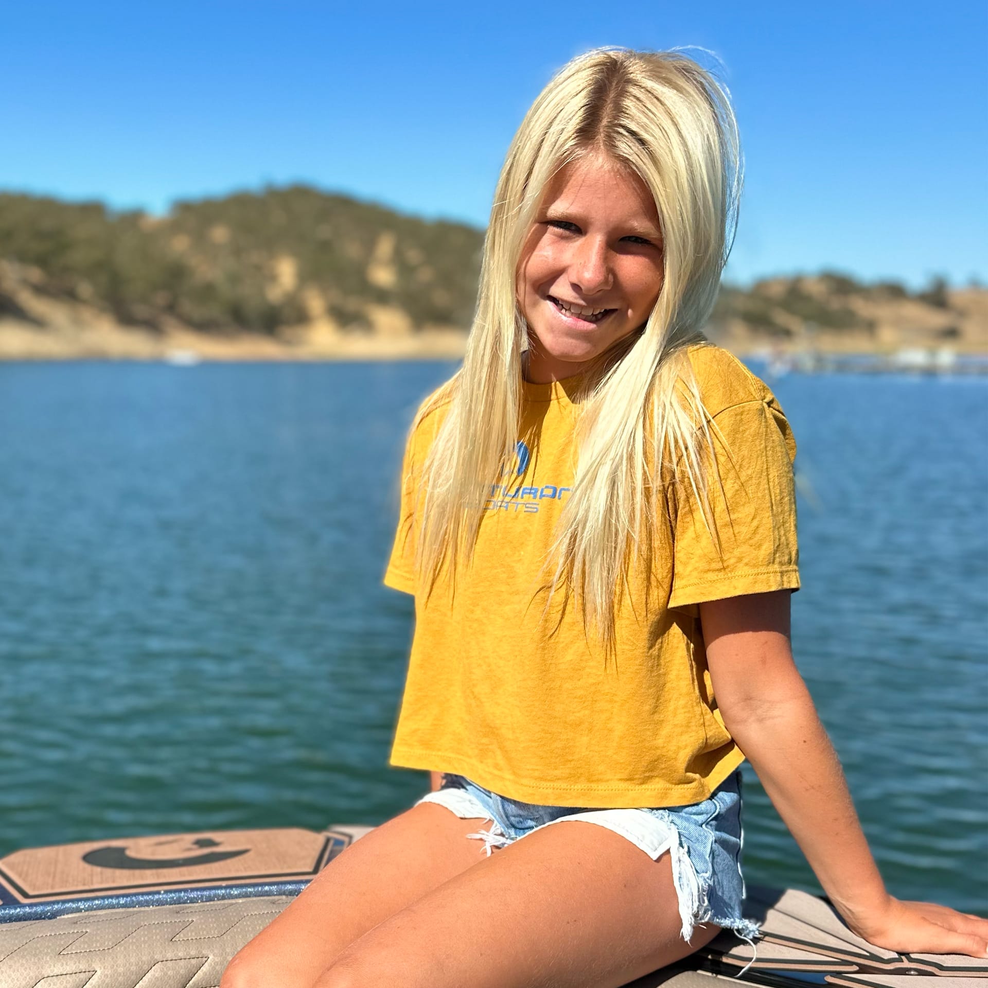 A person with long blonde hair, wearing a yellow shirt and denim shorts, sitting on a dock by a large body of water with a hilly landscape in the background.