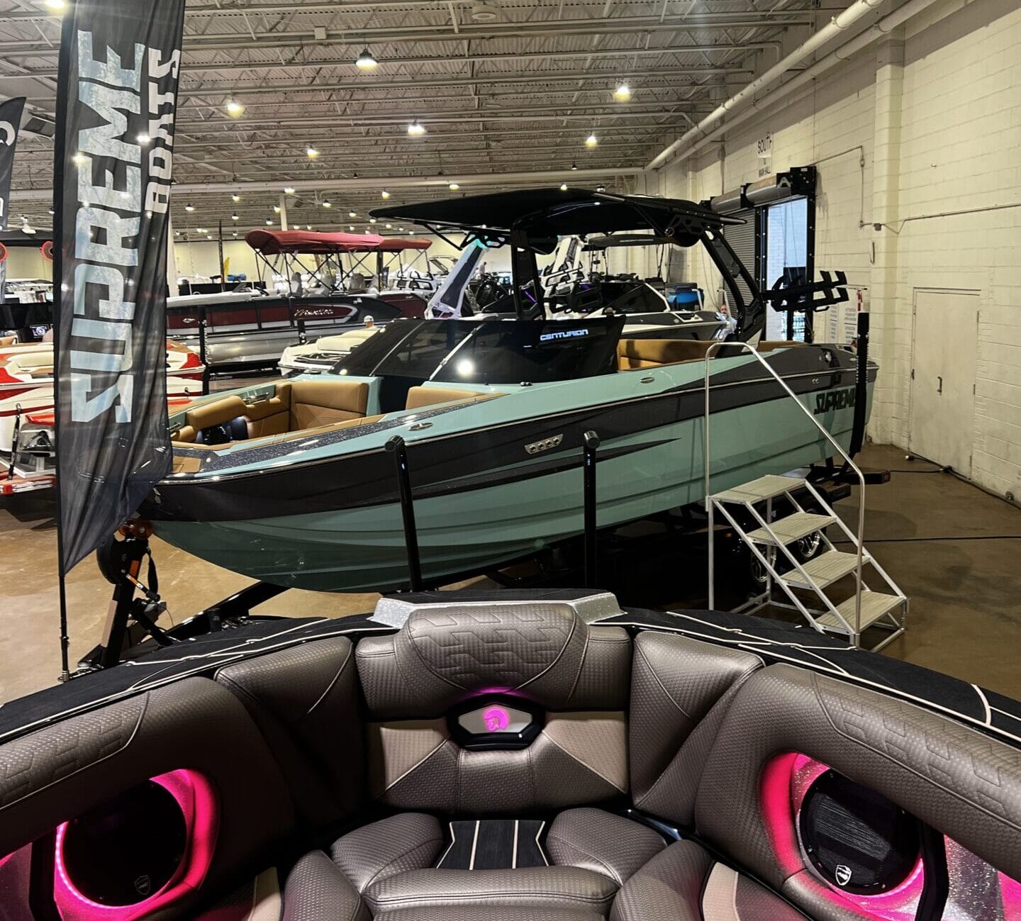 A boat showroom with various models, prominently featuring a green and black watercraft labeled "Supreme Boats". The foreground shows part of a boat interior with pink-lit cup holders.