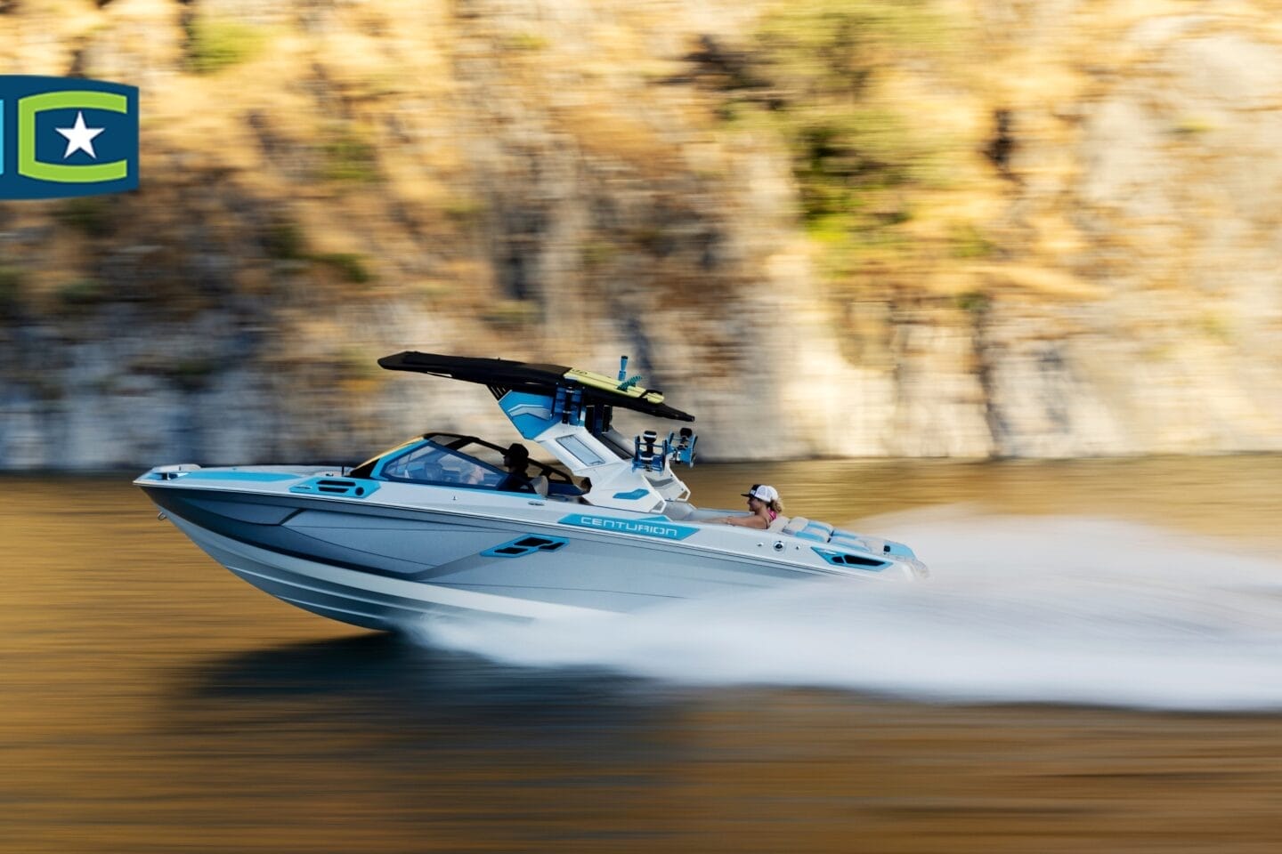 A speedboat with a wakeboard tower quickly travels on calm water near a rocky shore. A logo with a star is seen in the upper left corner.