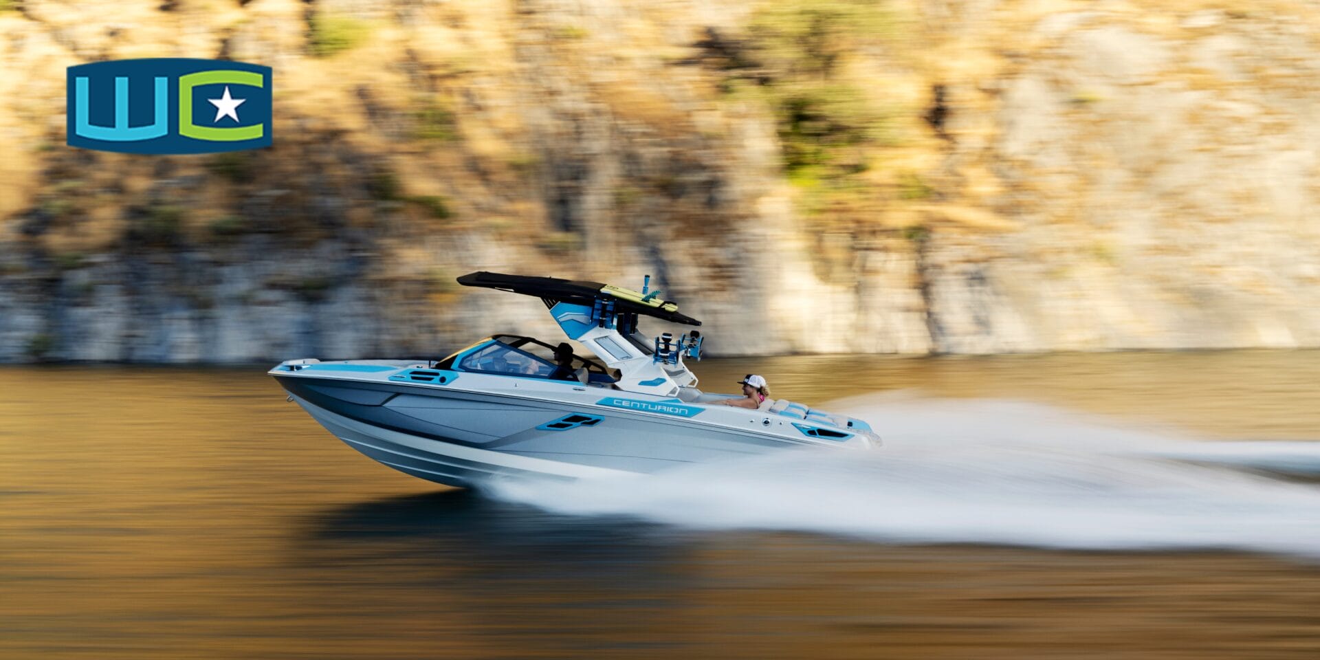 A speedboat with a wakeboard tower quickly travels on calm water near a rocky shore. A logo with a star is seen in the upper left corner.