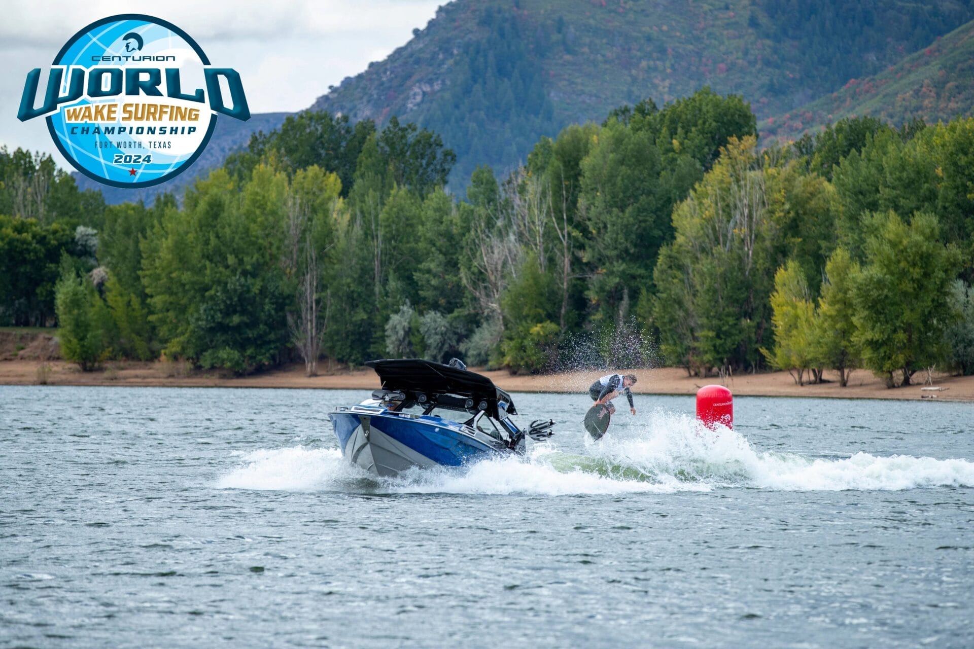 A person performs a wake surfing trick behind a speedboat on a lake with a forested hillside in the background. A logo indicates 