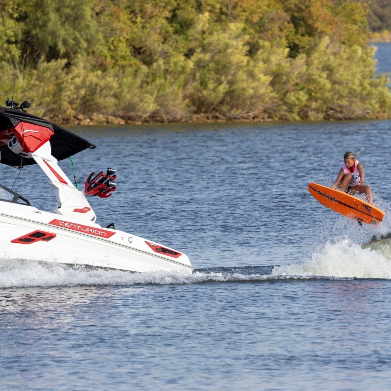 A person on an orange wakeboard, reminiscent of a Centurion Wake Surfer, performs a jump in the wake behind a white motorboat on a lake, with green shrubbery in the background. It's as thrilling as watching Brynlee Hall conquer waves with style.