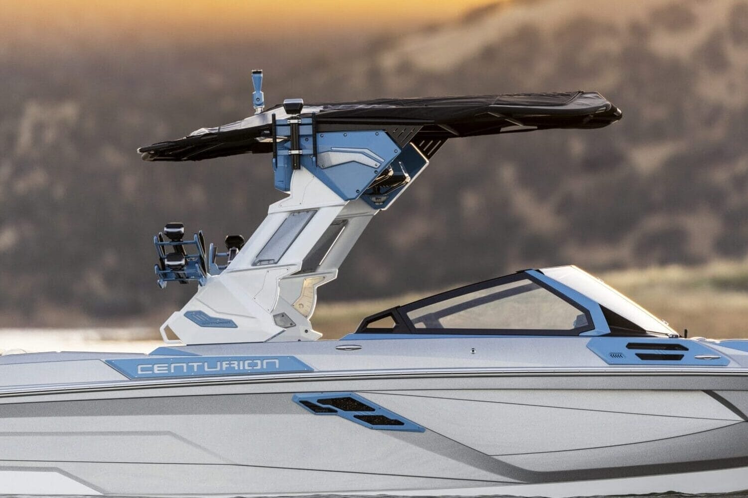 A sleek, silver and blue Centurion speedboat with the innovative Opti-V Hull glides effortlessly on calm water, its black canopy casting a serene silhouette against a backdrop of rolling hills at sunset.