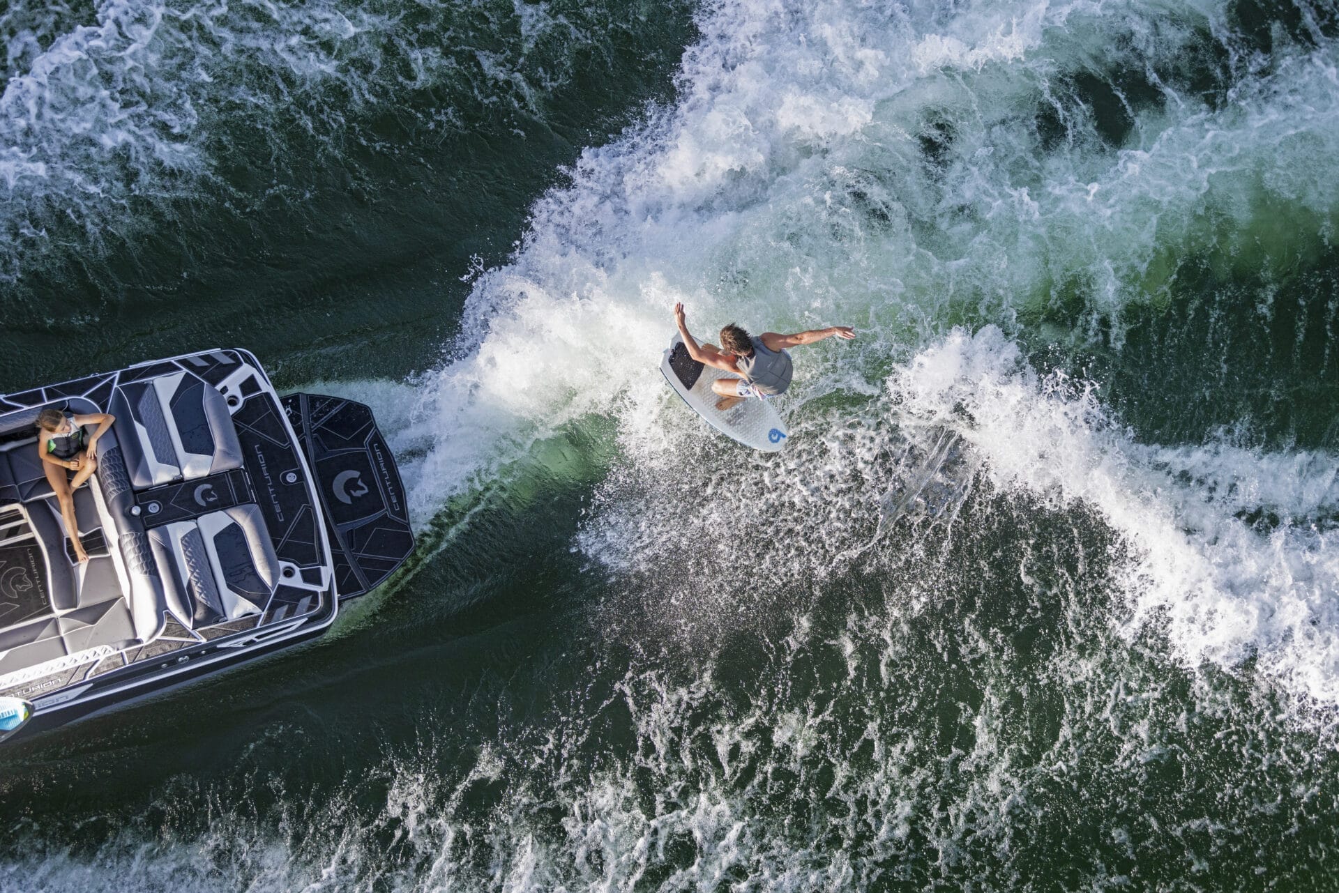 A person surfs the wake behind a speedboat on a green body of water.