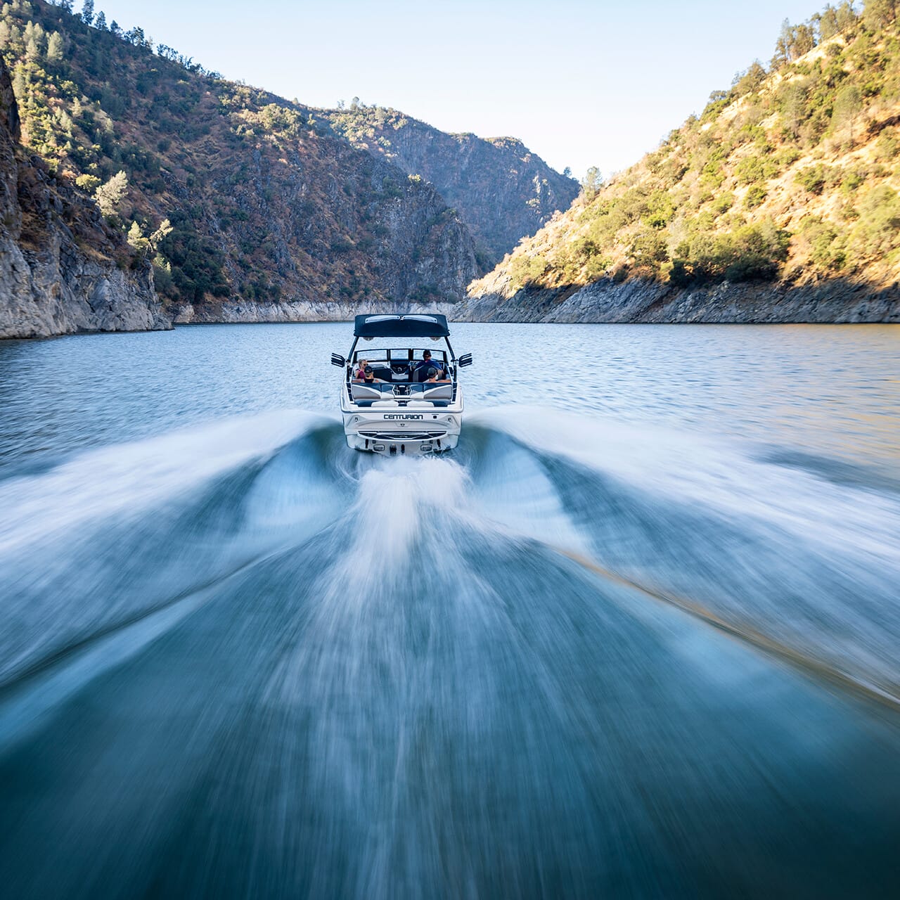 A Centurion Fe Series speedboat travels swiftly through a narrow canyon waterway, creating a wide, foamy wake. Lush, tree-covered hills rise steeply on either side under a clear blue sky.