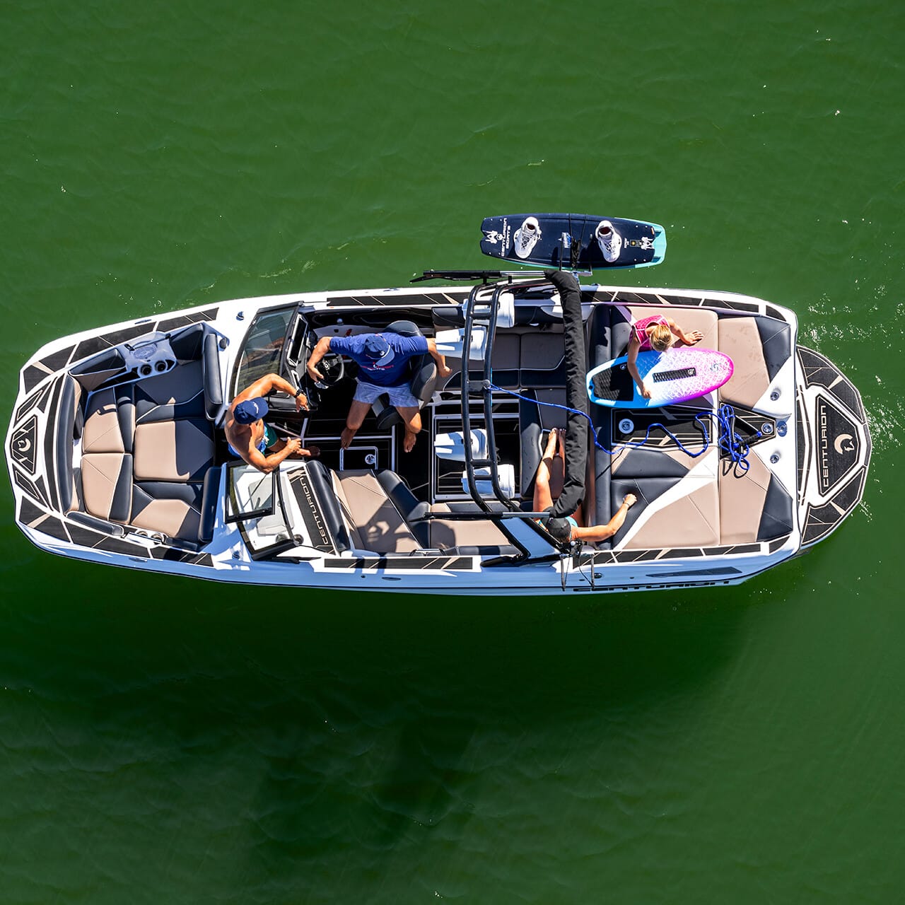 Aerial view of three people on a Centurion Fe Series boat gliding through green water, with two surfboards on board.