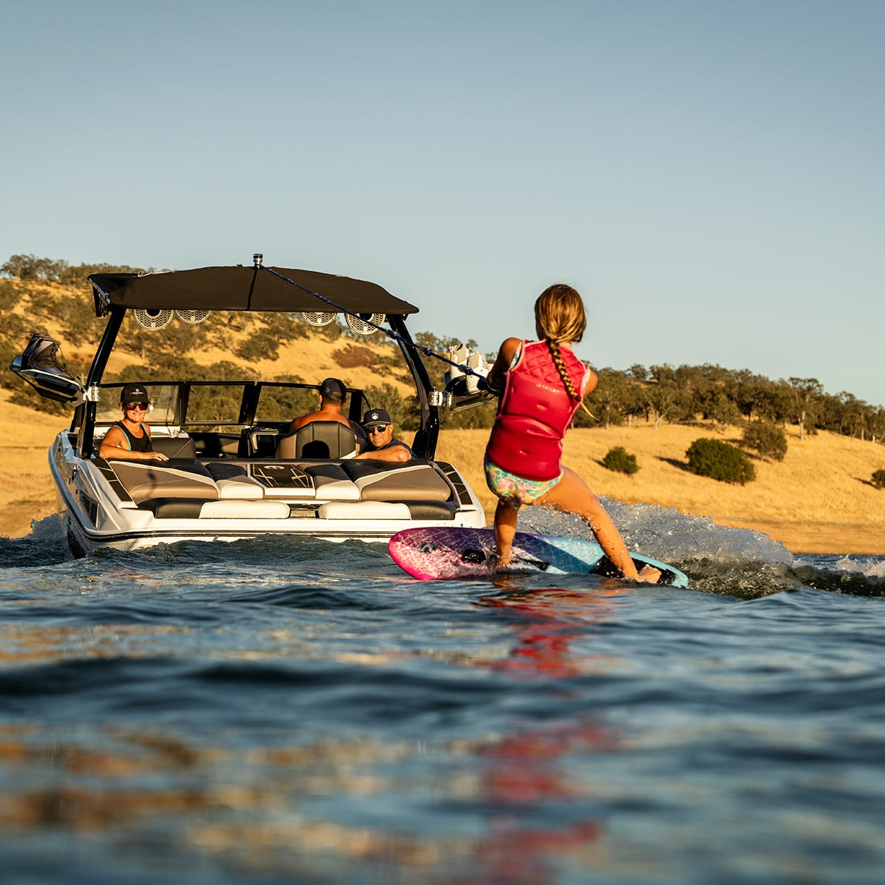 A person in a life vest wakeboards on a lake behind a sleek Centurion Fe Series motorboat under a clear sky, with passengers observing from the boat. Hills roll majestically in the background.