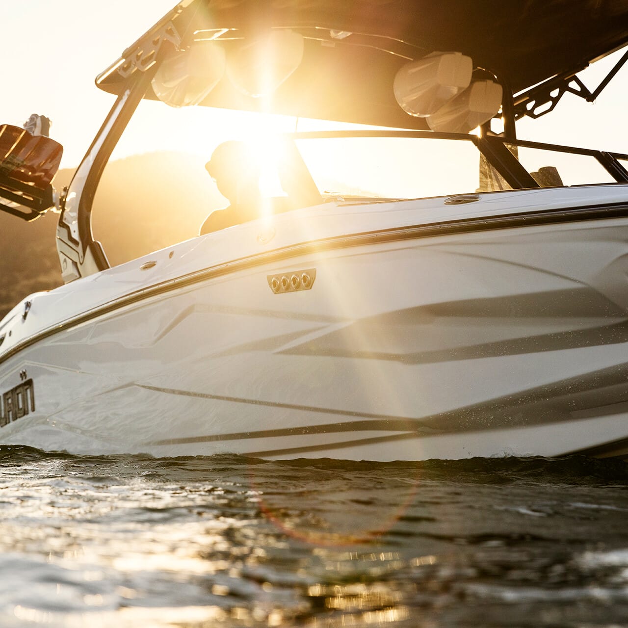 A sleek Centurion Fe Series boat glides on the water, with a person partially visible under the sunlit canopy. Hills form a serene backdrop, enhancing the picturesque scene.