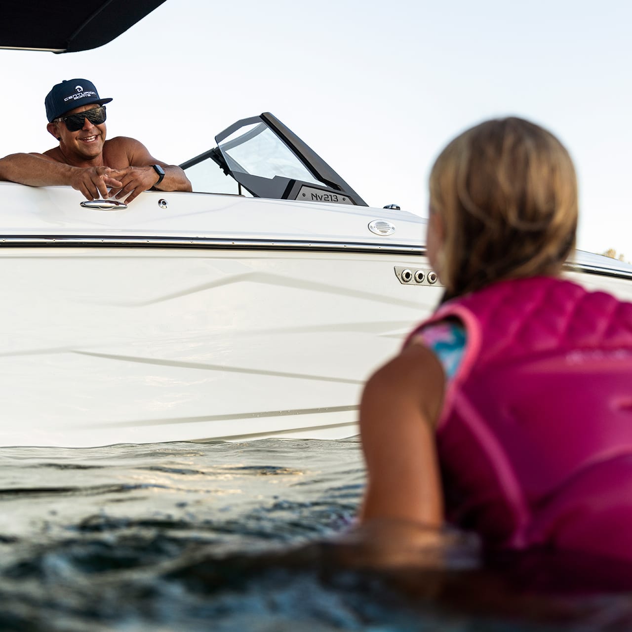 A person in a pink life jacket swims near a white Centurion Fe Series boat, chatting with someone leaning out.