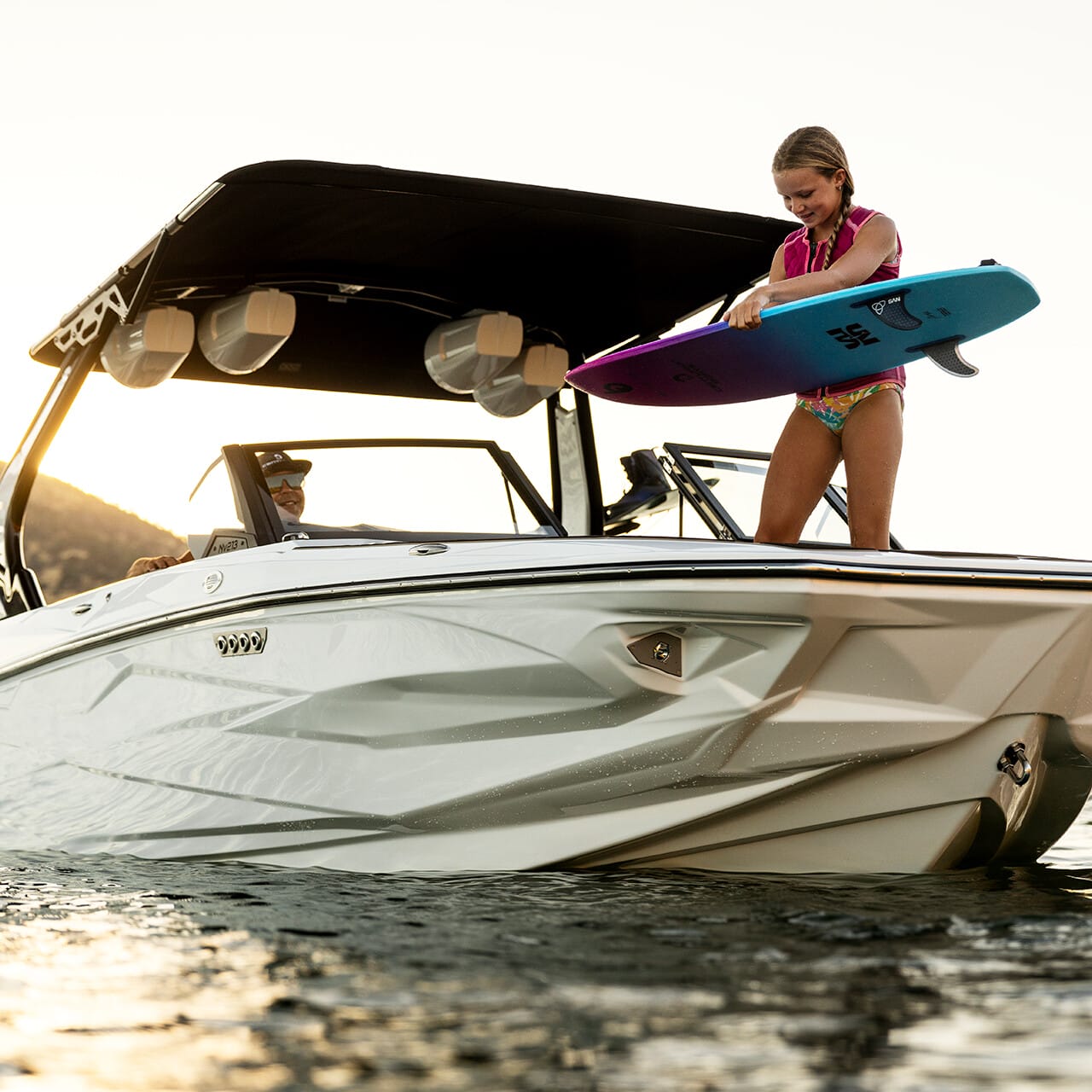 A person stands on a sleek white boat in the water, holding a colorful surfboard under a clear sky. The Centurion Fe Series vessel embodies both style and performance, promising an unforgettable adventure on the open water.