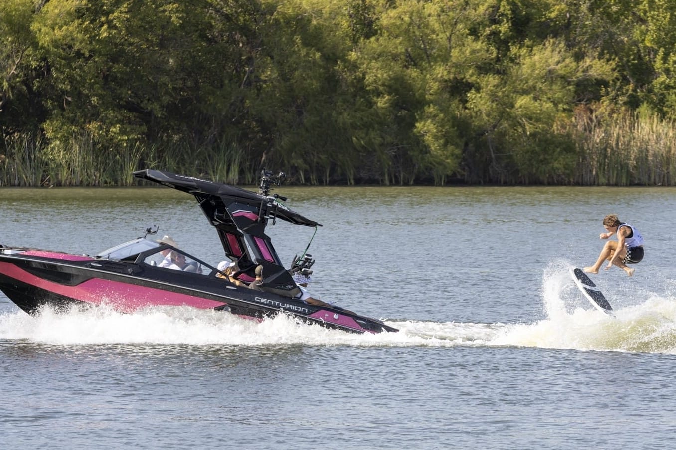A person is wakeboarding behind a speedboat on a lake with trees in the background.