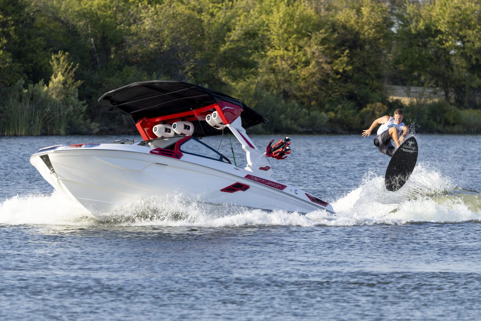 A person wakeboarding performs a trick in the air beside a moving motorboat on a lake with trees in the background.