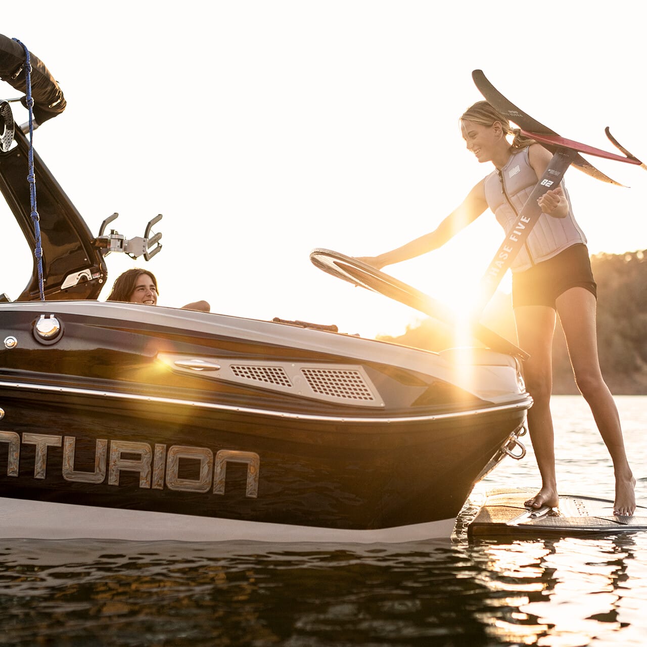 A person on a boat reaches out to help a woman with water skis step onto the boat at sunset.