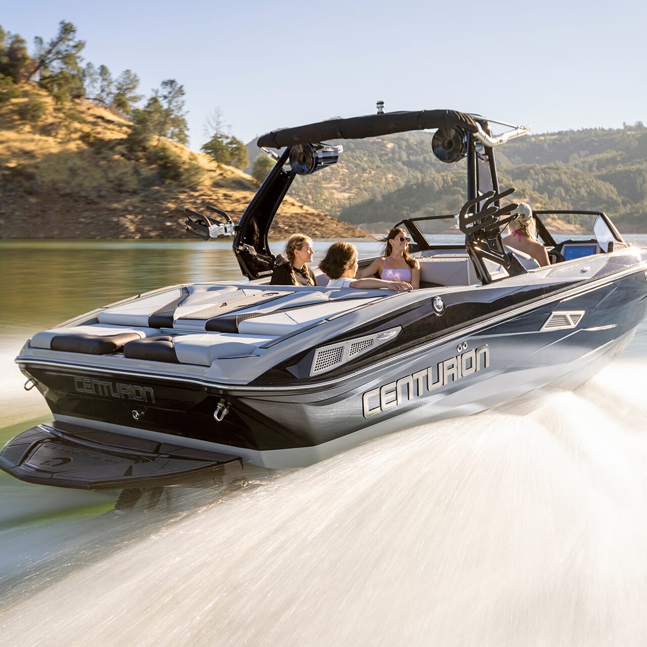 A sleek motorboat with four people on board speeds across a calm lake, surrounded by green landscape.