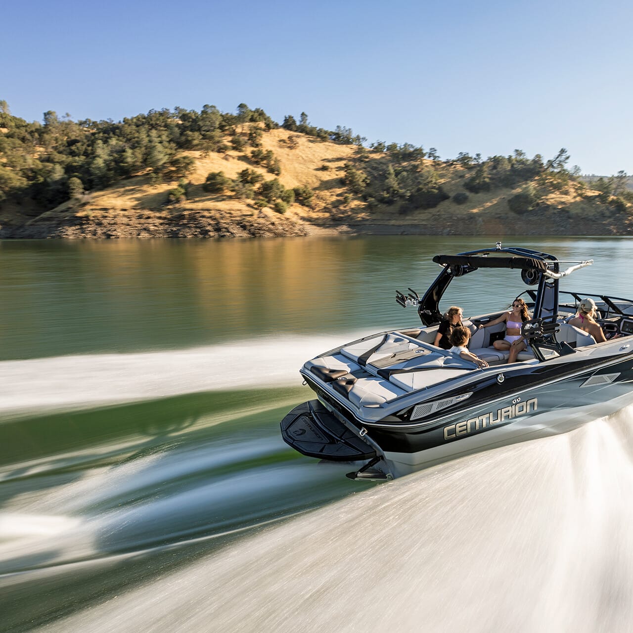 A speedboat with several passengers glides swiftly across a lake, creating a trail on the water, with tree-covered hills in the background.