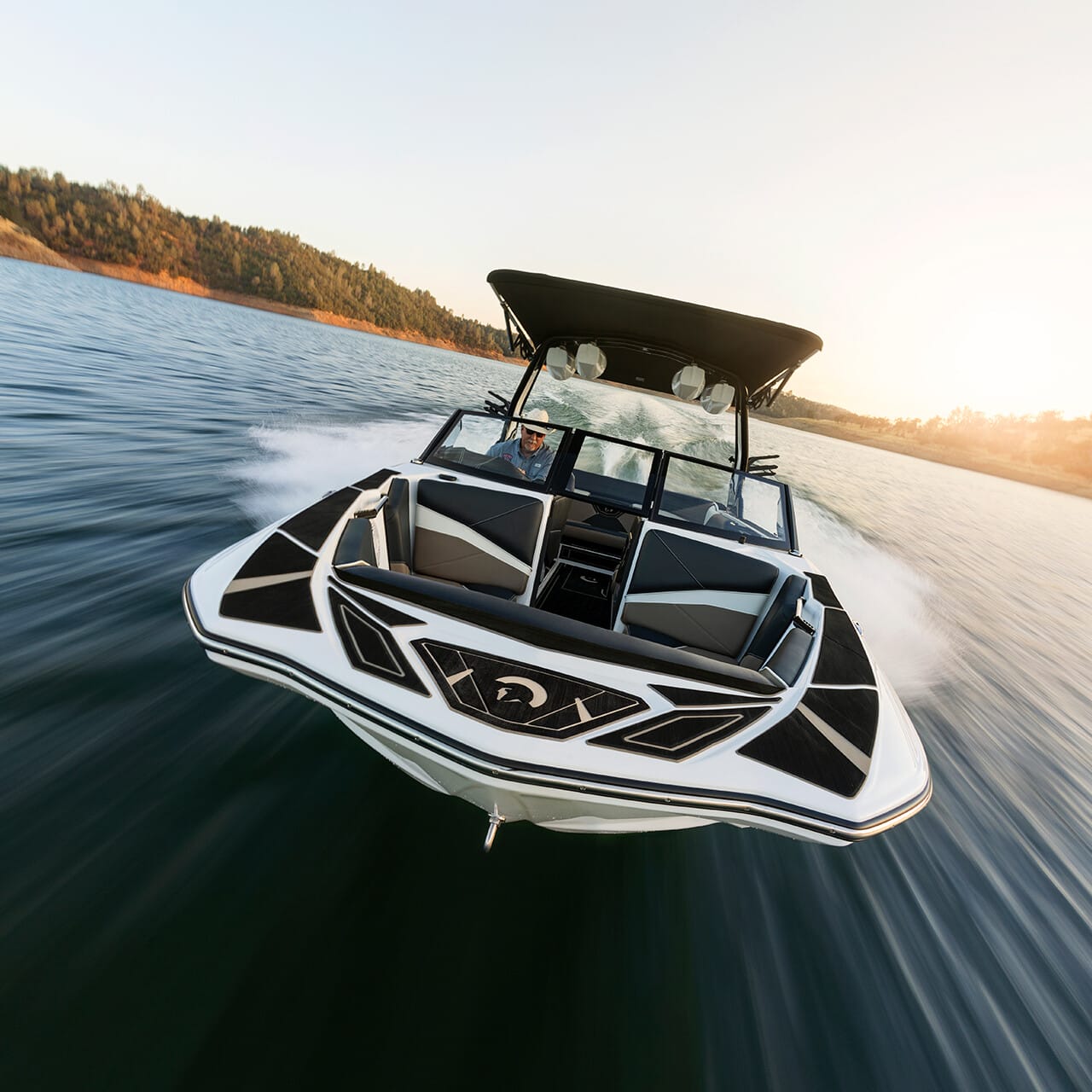 A speedboat glides swiftly on a lake with water rippling behind, against a backdrop of trees and a setting sun.