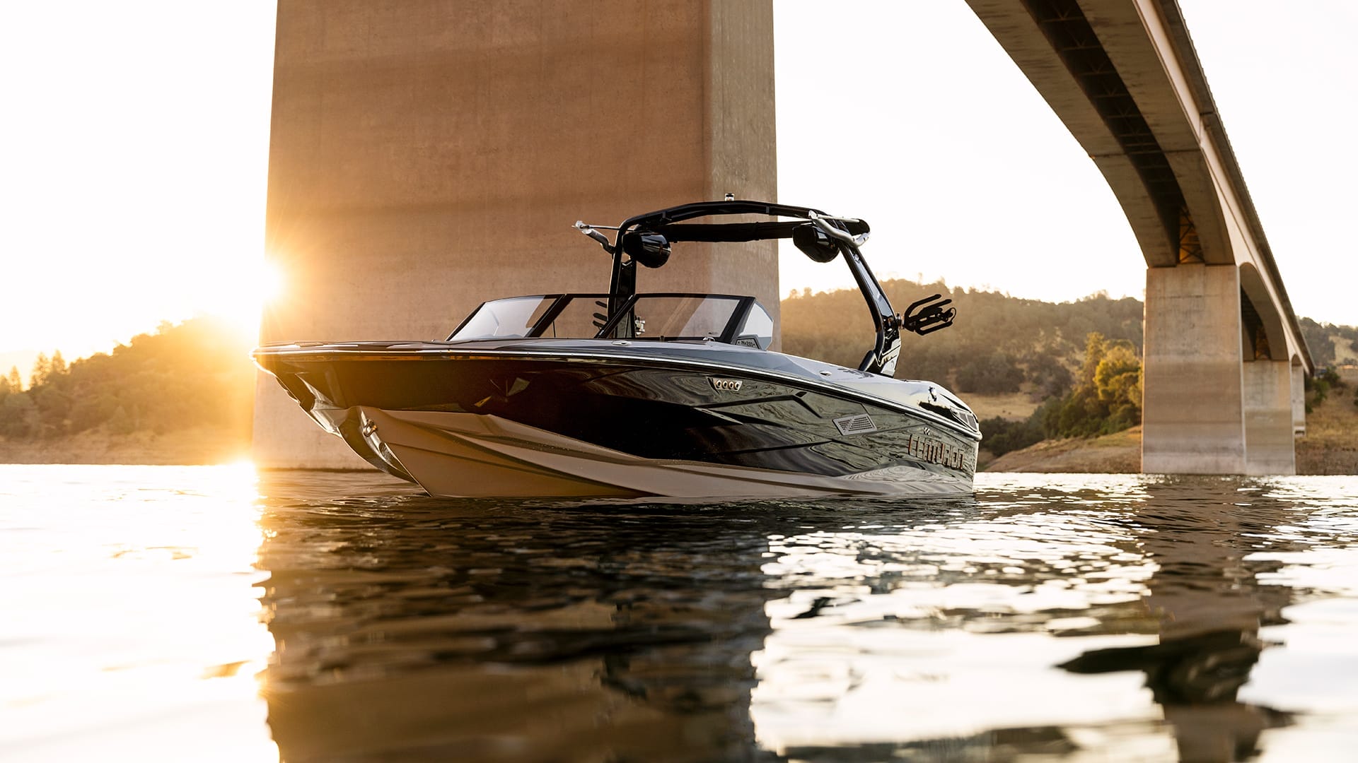 A sleek motorboat floats on calm water beneath a large bridge at sunset.