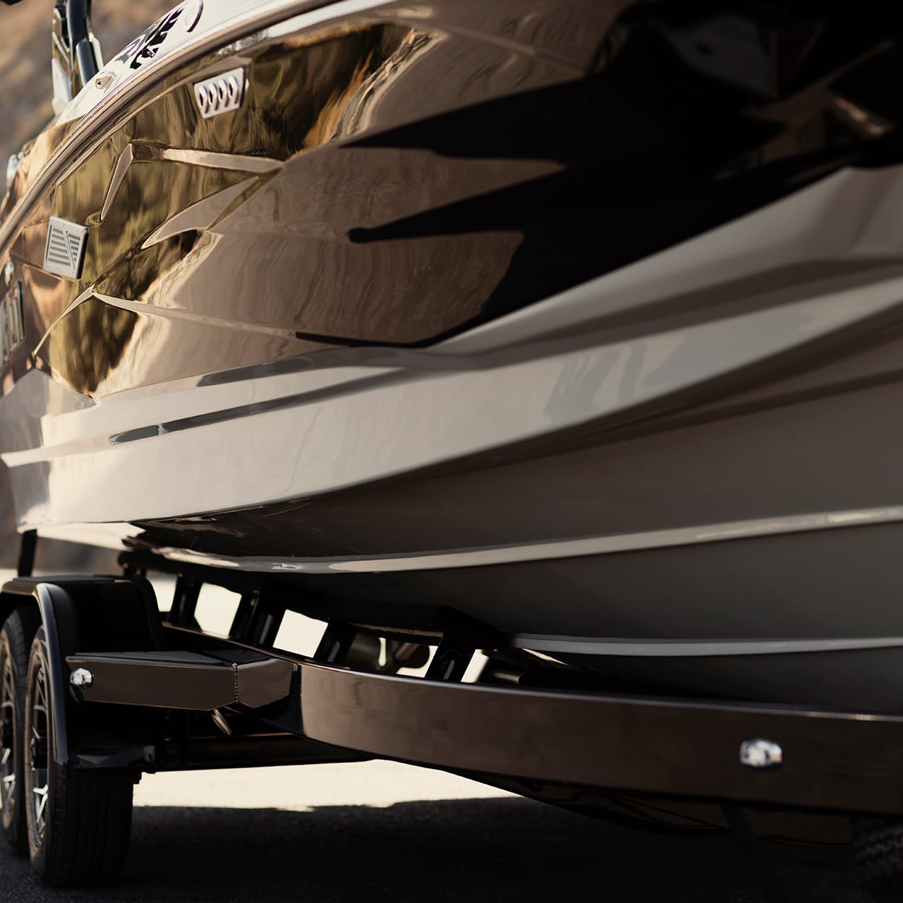 A close-up view of a boat's hull mounted on a trailer, reflecting sunlight, with rocky terrain in the background.